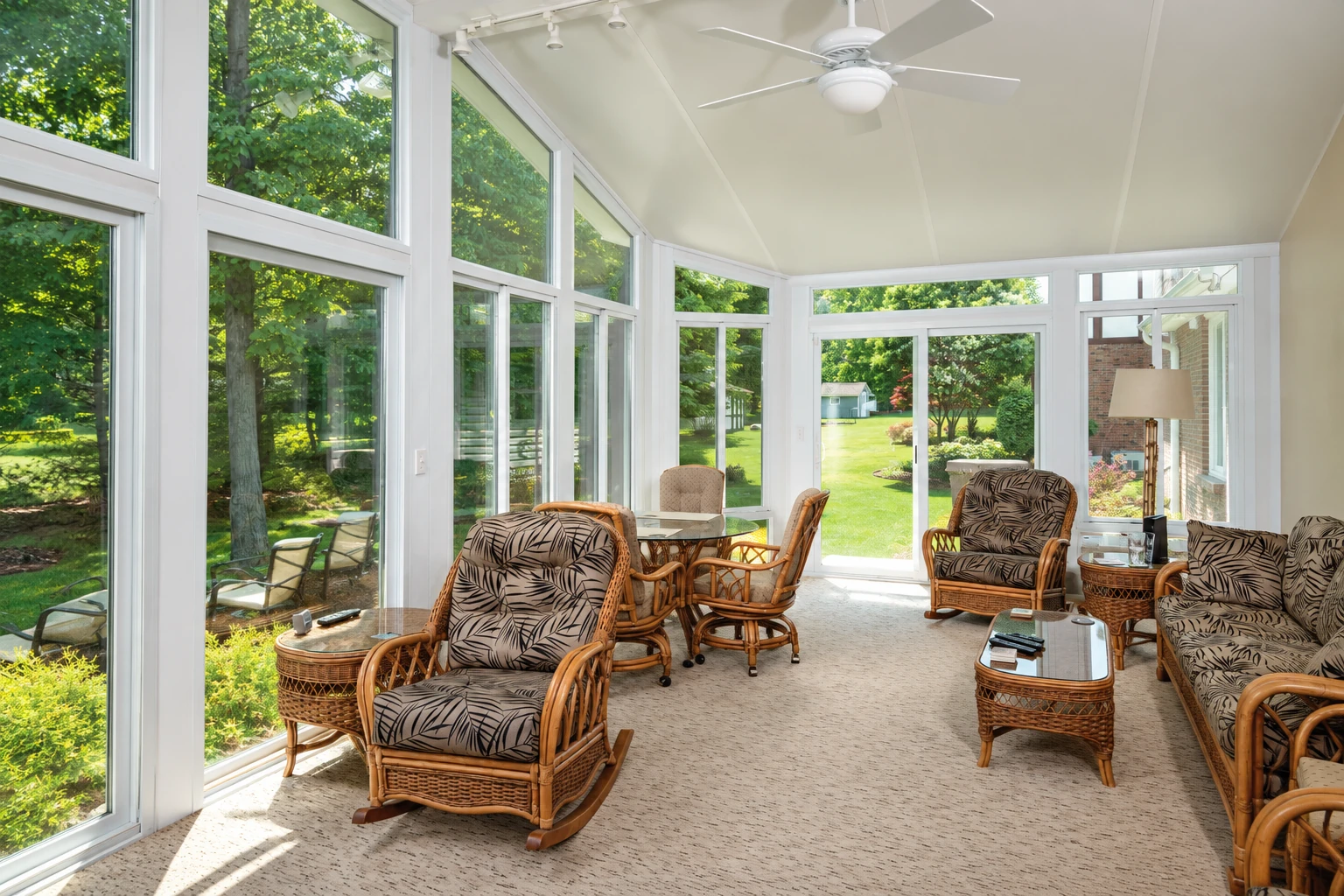 Four-season sunroom with white-framed windows, a ceiling fan, and wicker chairs overlooking a leafy garden.