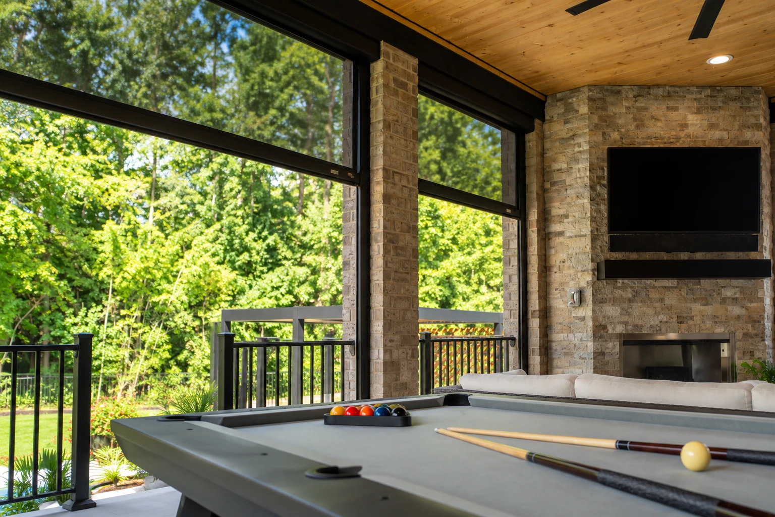 Covered patio with a wood-paneled ceiling, stone fireplace wall, and a pool table beside open views of trees.