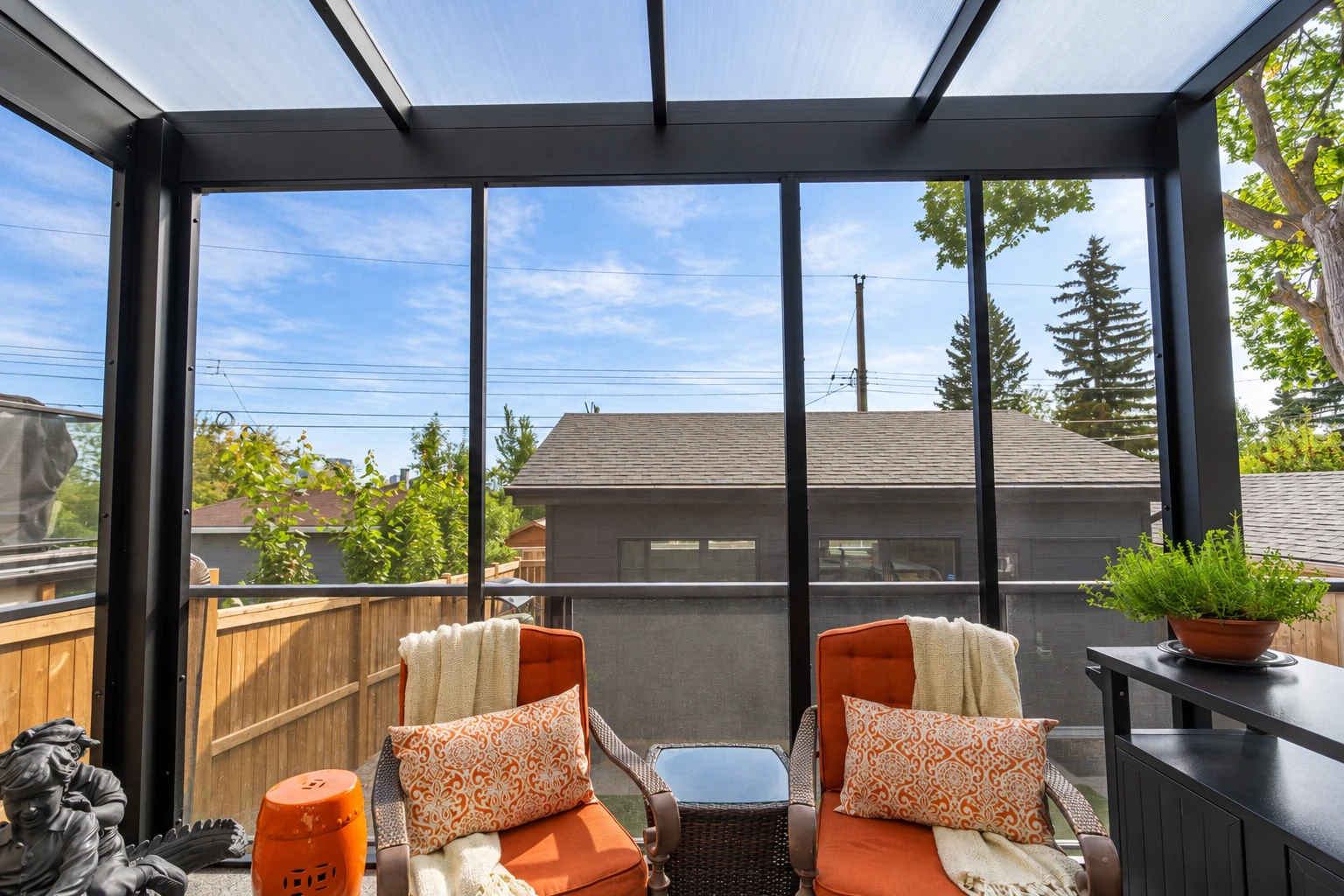 Enclosed patio with a dark frame and translucent roof, orange-cushioned chairs, and a view of neighboring homes.