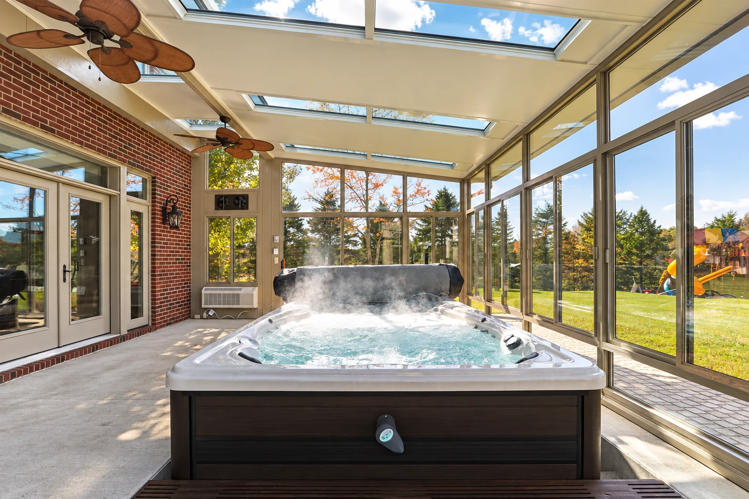 Glass solarium with a bubbling hot tub, skylights, ceiling fans, and views of a leafy yard.