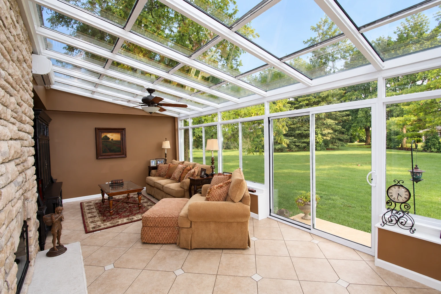 Solarium with a glass-paneled roof and walls, tile flooring, and tan sofas facing a green lawn.