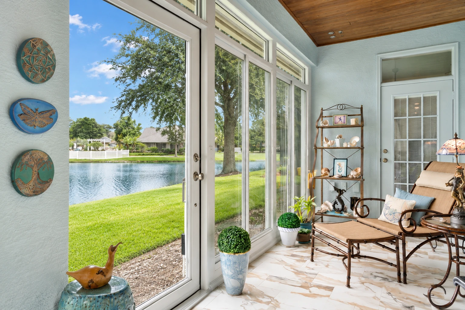 Sunroom addition with tall white-framed windows overlooking a pond, furnished with a wicker seating set and potted plants.