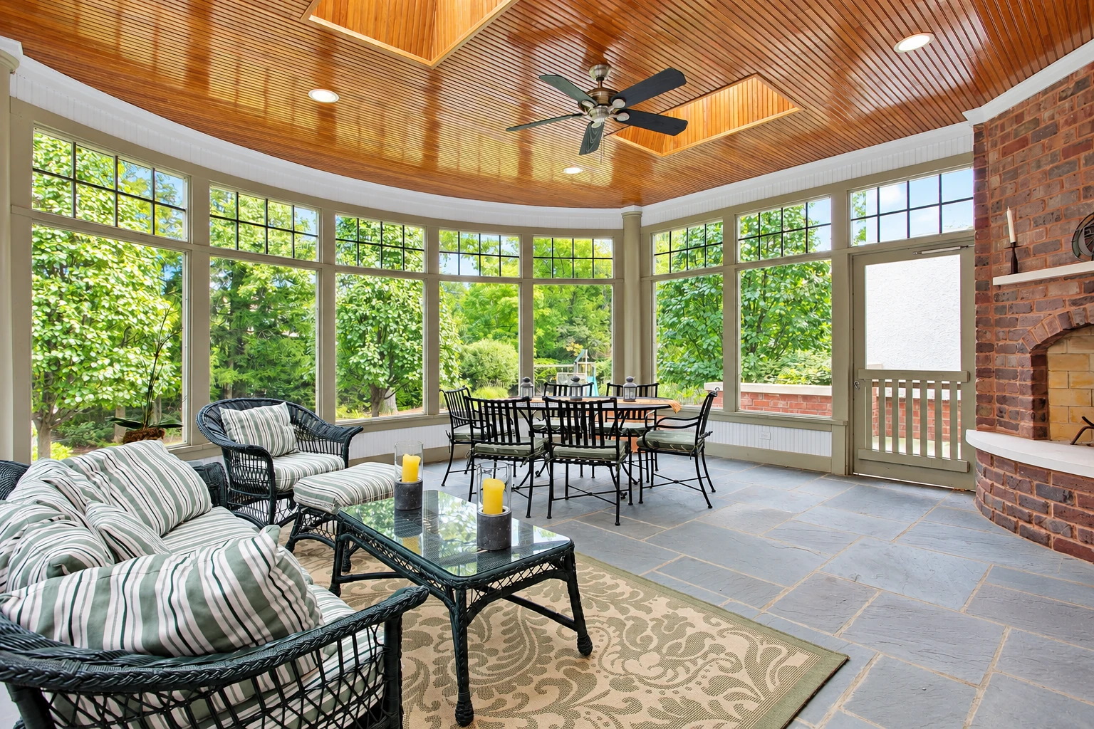 Sunroom addition with a wood-paneled ceiling, large windows, brick fireplace, dining set, and striped wicker seating.