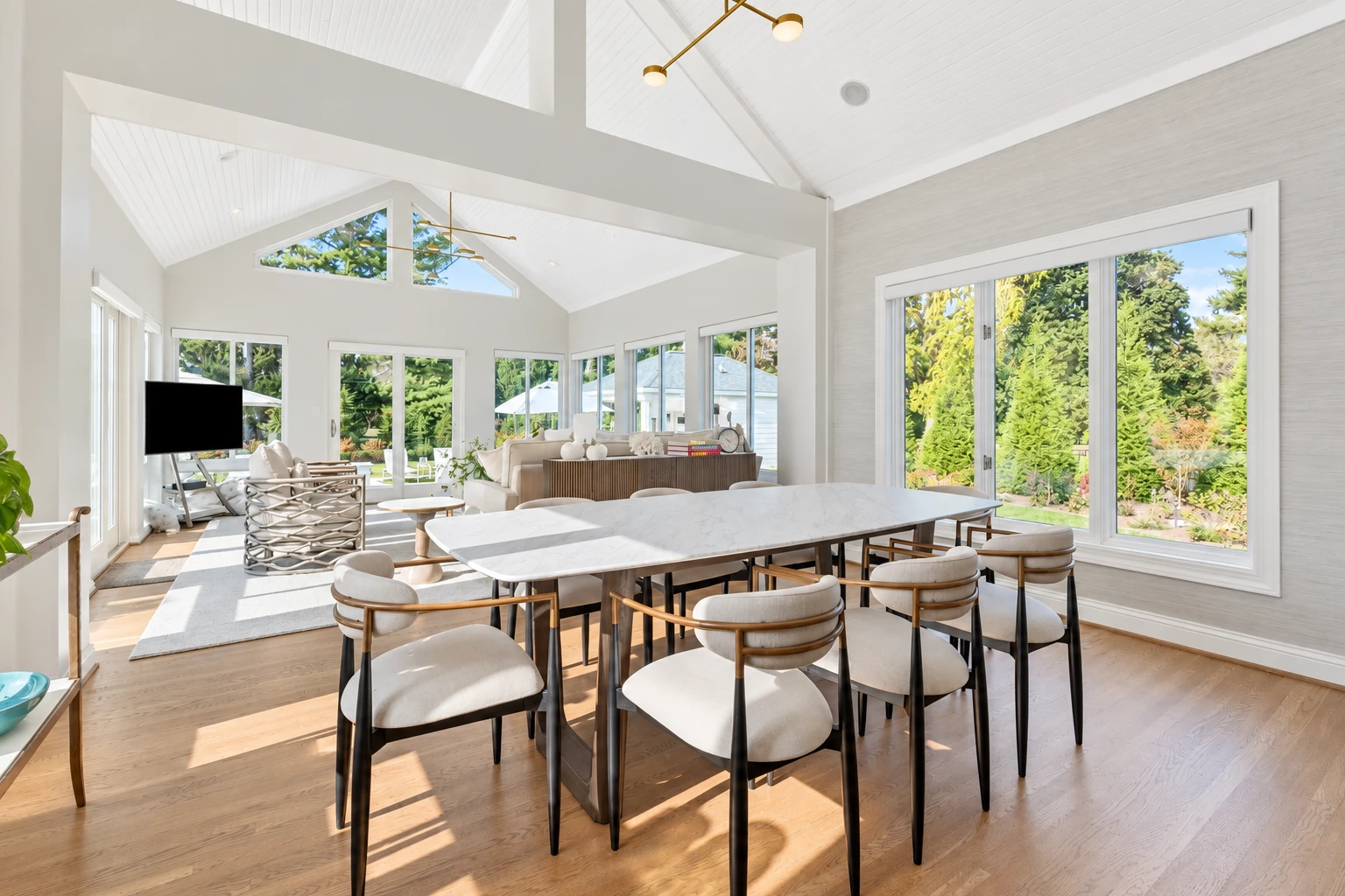 Bright sunroom with a vaulted white ceiling, a large marble dining table, and windows overlooking a leafy garden.