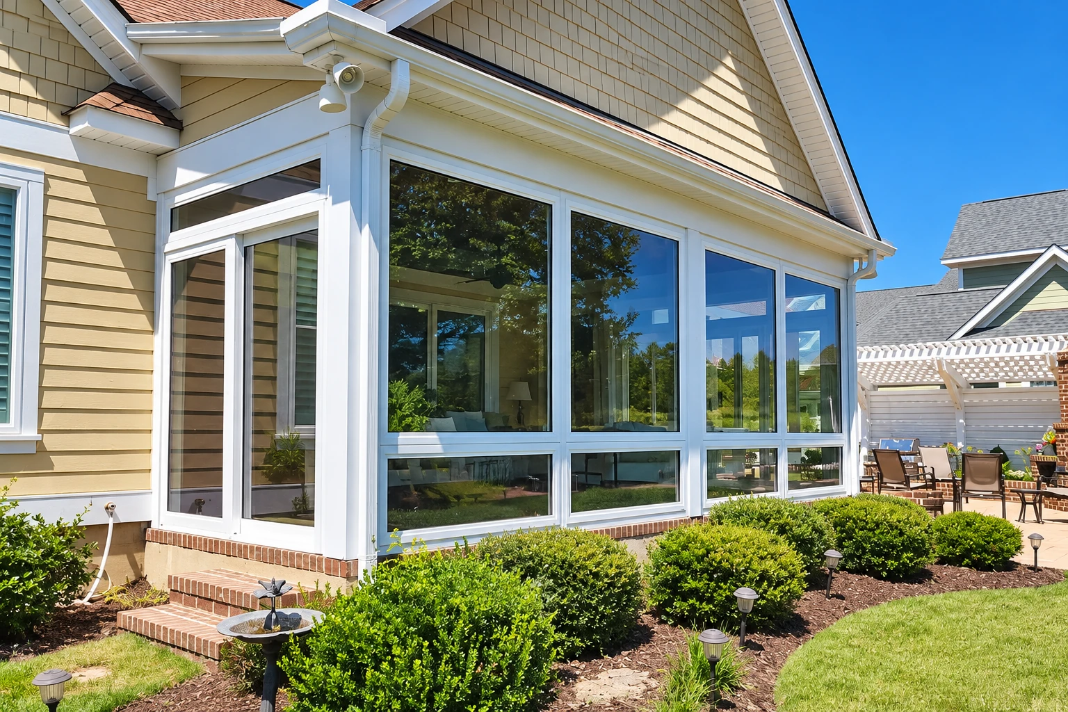 White-framed sunroom addition on a tan-sided house, with broad windows, a glass door and landscaped garden beds.