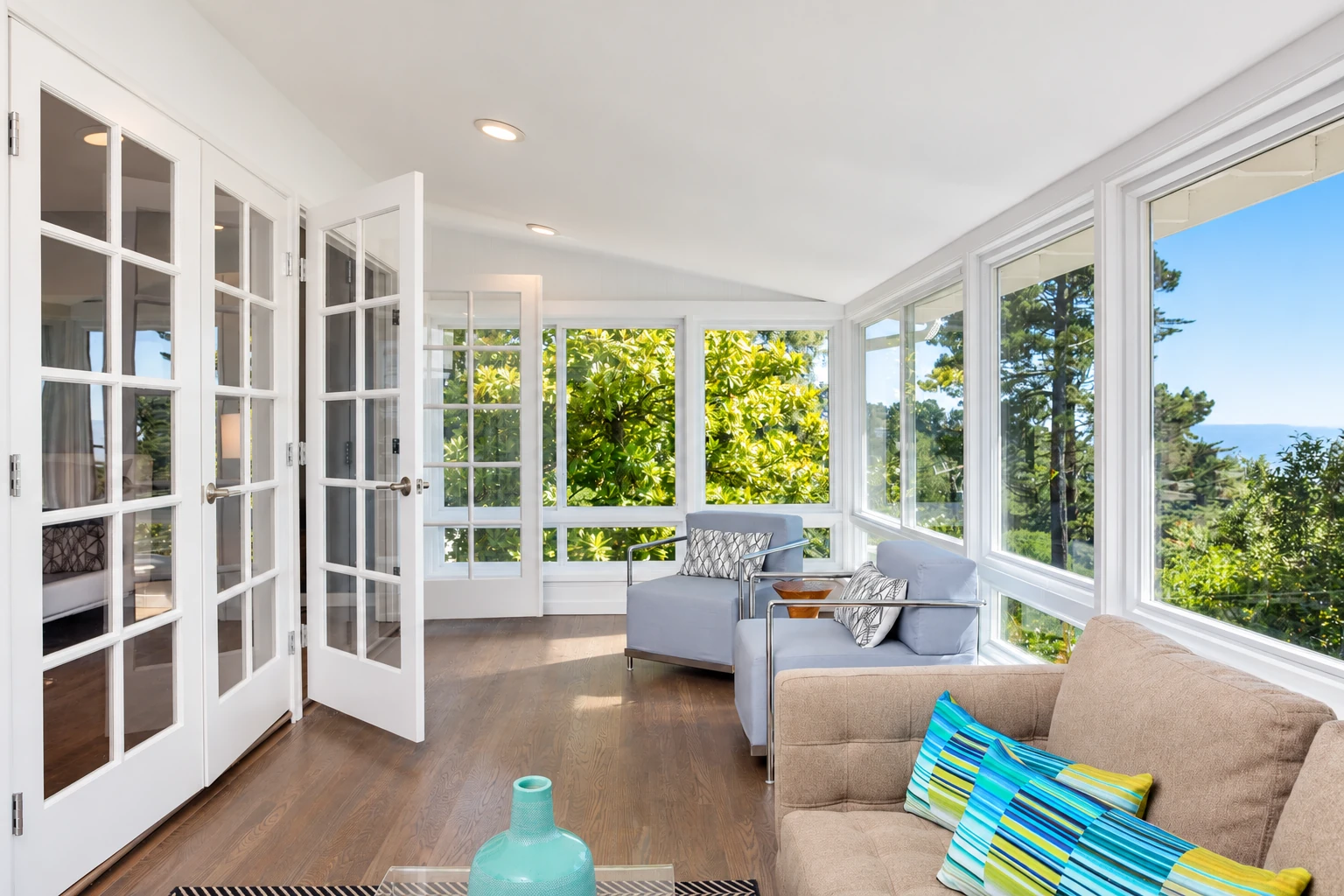 Sunroom with white-framed windows, open French doors, wood flooring, and pale blue and beige seating.
