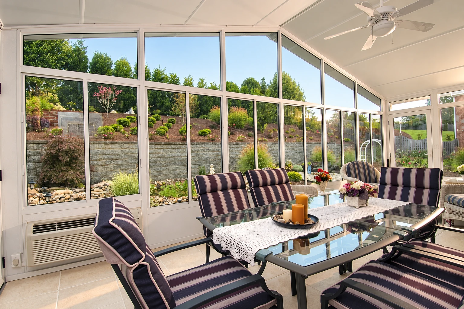 Enclosed sunroom with tall windows, striped lounge chairs, and a glass table facing a landscaped garden.