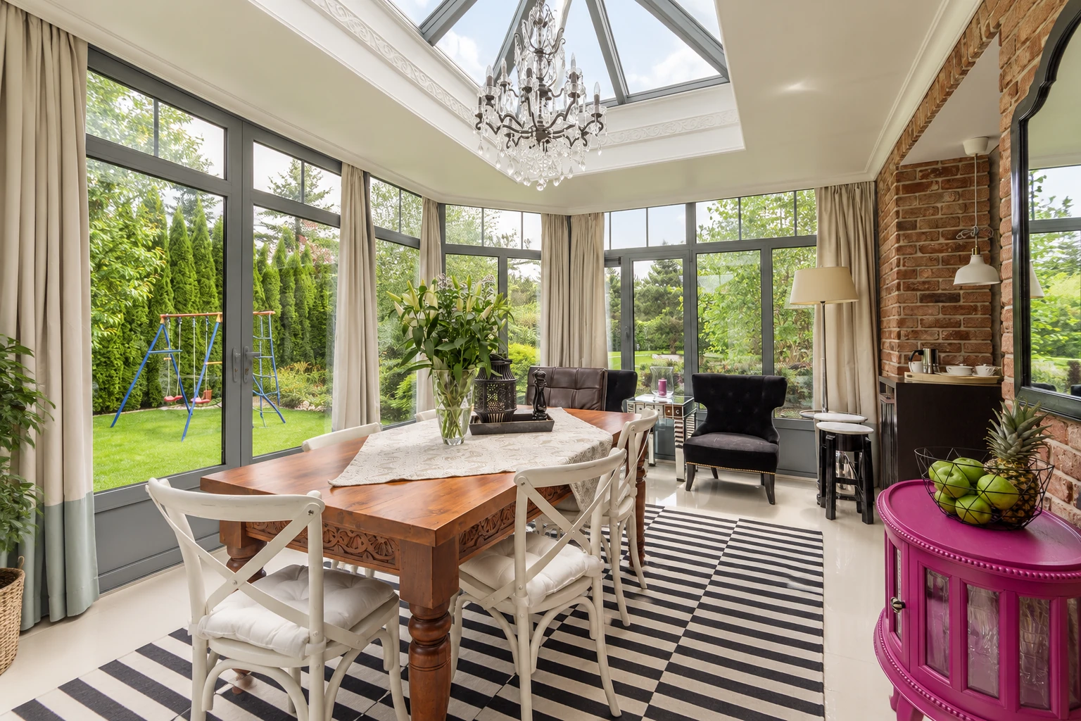Bright sunroom dining area with a skylight, chandelier, large windows, and a wooden table on a striped rug.